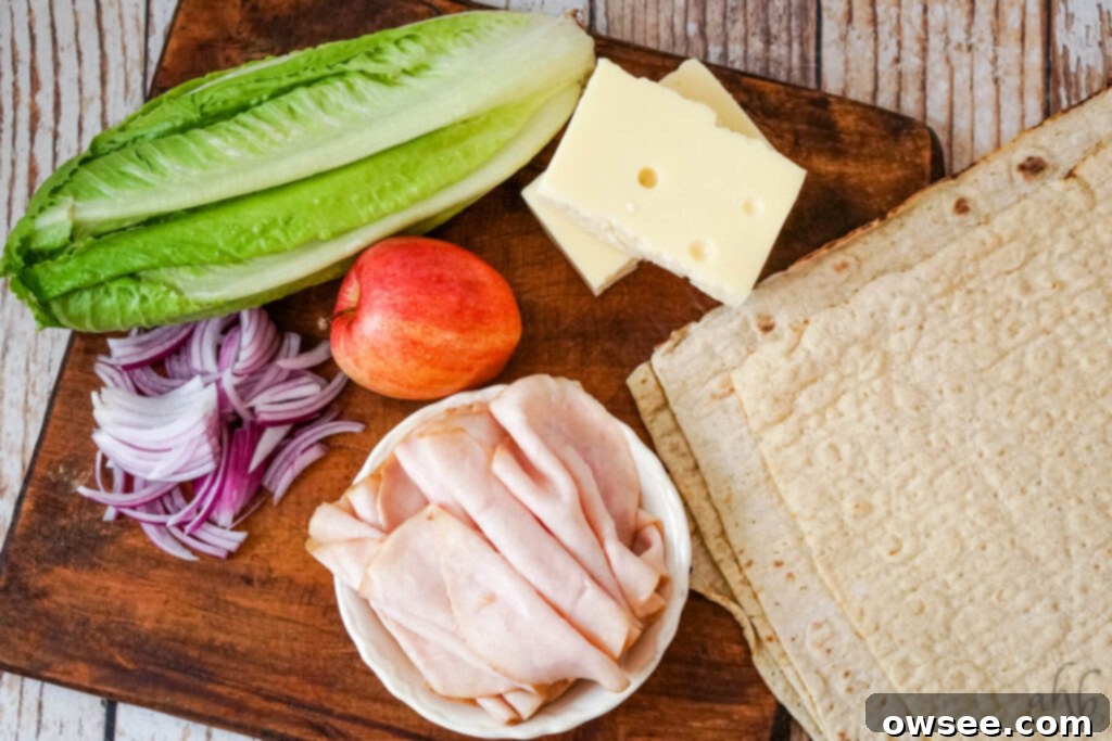 Crisp Turkey Apple Swiss Wraps 5 A head of romaine lettuce, a red apple, two blocks of swiss cheese, thinly sliced red onion, sliced turkey, and four large tortillas on a wooden cutting board.