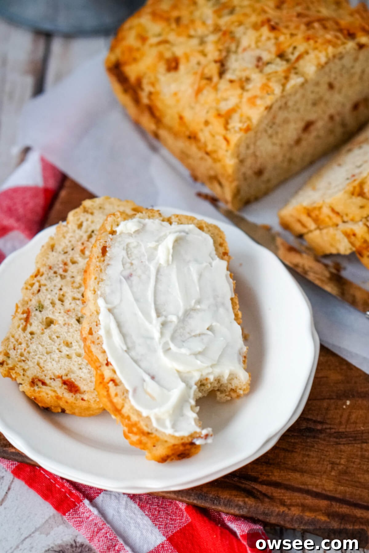 Sun-Kissed Tomato Parmesan Beer Bread 3 Slices of Sun-dried Tomato and Parmesan Beer Bread, with two slices served on a white plate alongside a dollop of cream cheese, one slice having a bite taken.