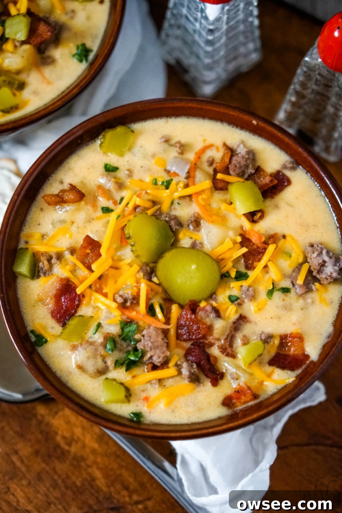 Creamy Slow Cooker Bacon Cheeseburger Soup 10 A bowl of Slow Cooker Bacon Cheeseburger Soup served with shredded cheddar cheese, sliced pickles, and bacon pieces.