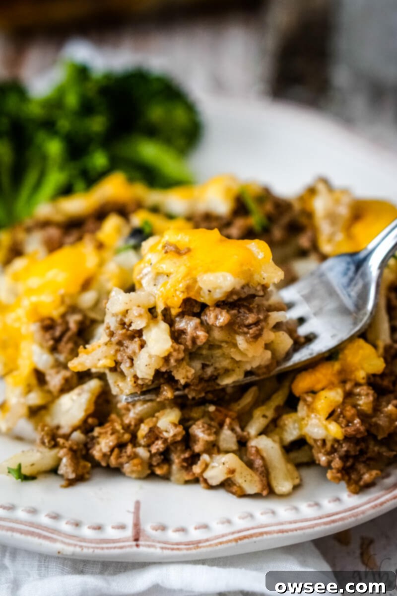 Savory Beef and Hashbrown Bake 3 A forkful of Cheesy Hamburger Hashbrown Casserole held above a serving on a white plate with vibrant steamed broccoli, showcasing its rich texture.