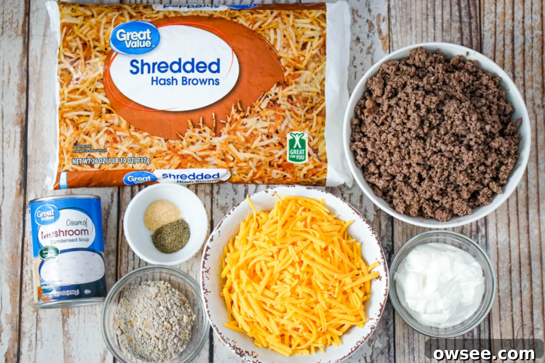 Savory Beef and Hashbrown Bake 5 A selection of raw ingredients for Hamburger Hashbrown Casserole: a bag of shredded hashbrowns, a bowl of browned ground beef, a bowl of shredded cheddar cheese, black pepper, garlic powder, a can of cream of mushroom soup, dry onion soup mix, and a container of sour cream, all ready for preparation.