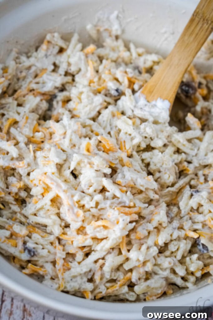 Savory Beef and Hashbrown Bake 7 Hashbrowns, shredded cheddar cheese, cream of mushroom soup, sour cream, black pepper, and garlic powder are being mixed thoroughly in a large white bowl with a wooden spoon for the cheesy potato layer.