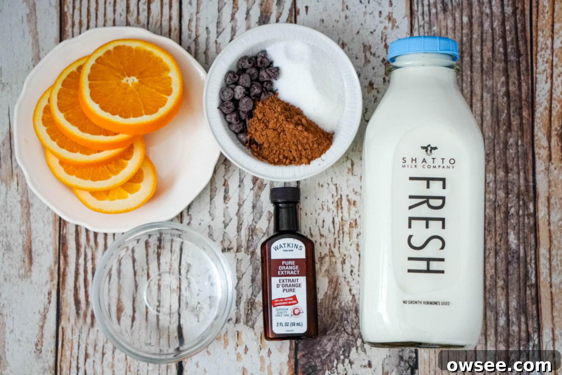 Zesty Cocoa Bliss 6 Slices of fresh orange on a white plate, alongside bowls containing granulated sugar, cocoa powder, and semi-sweet chocolate chips. Also pictured are a bowl of water, a bottle of orange extract, and a bottle of Shatto milk, all essential ingredients for Orange Hot Chocolate.