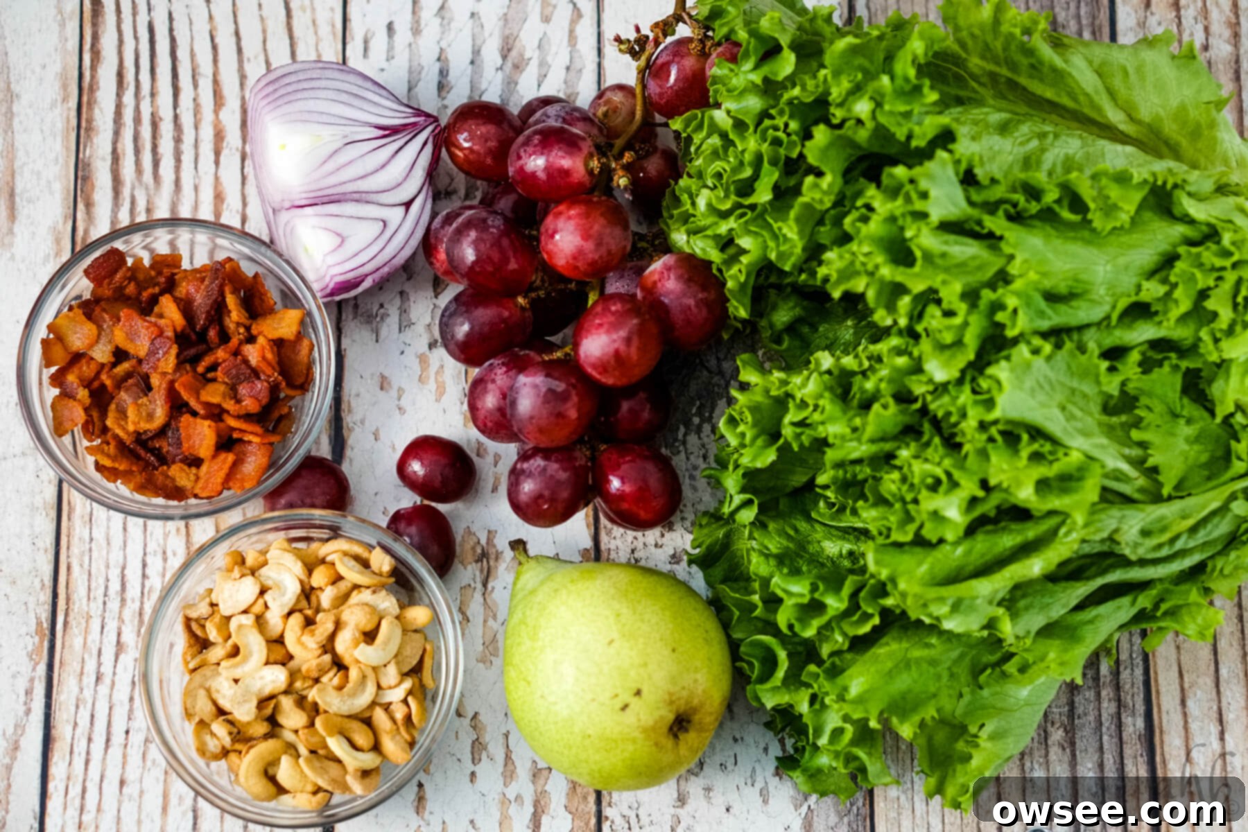 Cashew Salad 5 A head of lettuce, a pear, some red seedless grapes, half of a red onion, a bowl of bacon pieces and a bowl of cashew halves and pieces sit next to each other.