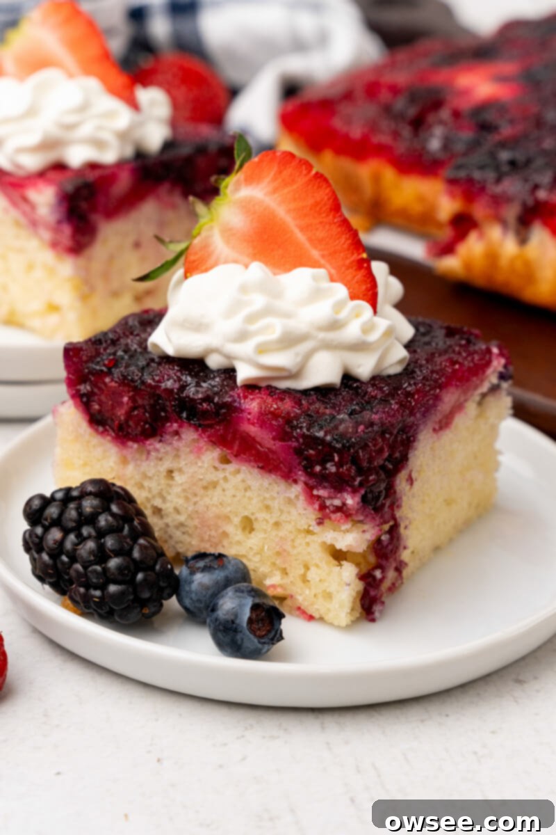 Caramelized Berry Flip Cake 2 A slice of Berry Upside Down Cake is served on a white plate, garnished with a fresh blackberry, two blueberries, a dollop of whipped cream, and half a strawberry.