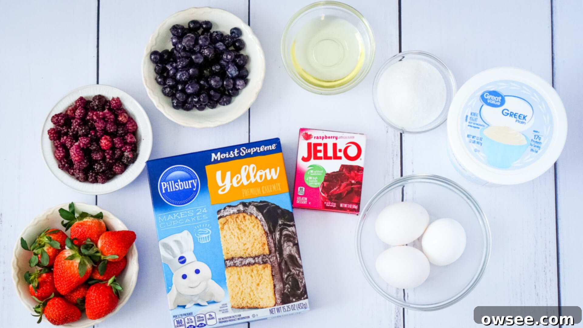 Caramelized Berry Flip Cake 5 Bowls containing fresh strawberries, blackberries, and blueberries, alongside a box of yellow cake mix, a box of raspberry gelatin, three eggs, vegetable oil, granulated sugar, and plain Greek yogurt, all arranged on a light surface.