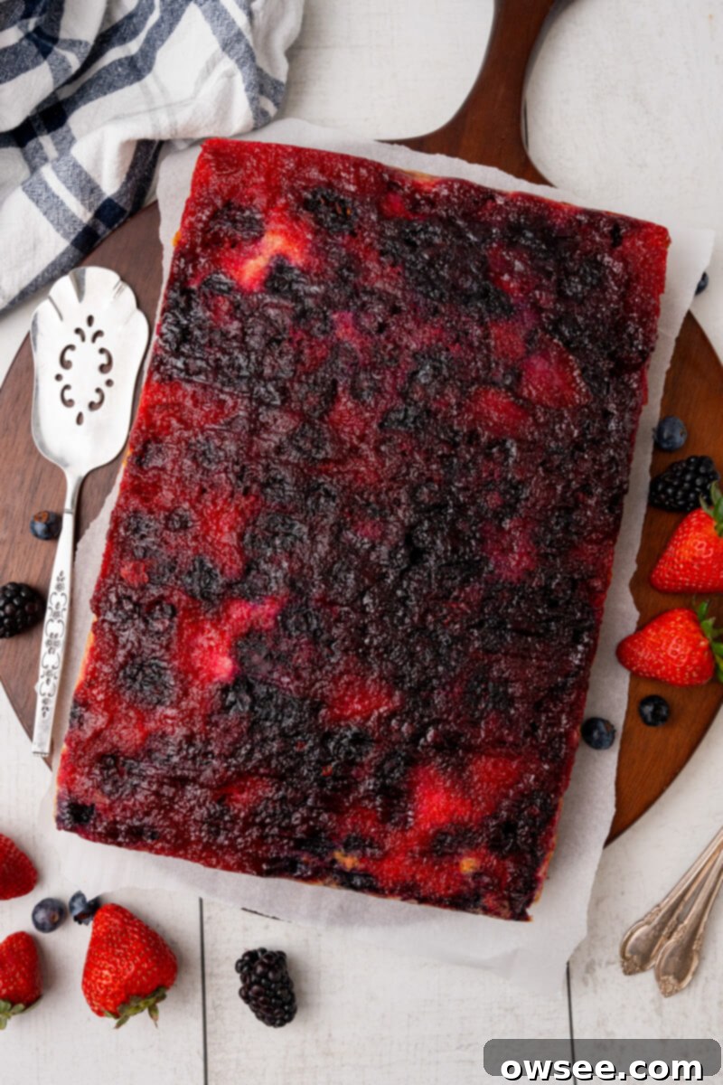 Caramelized Berry Flip Cake 9 A perfectly cooled Berry Upside Down Cake, vibrant with its berry topping, rests on a piece of parchment paper on a round serving platter, surrounded by extra fresh berries for garnish.
