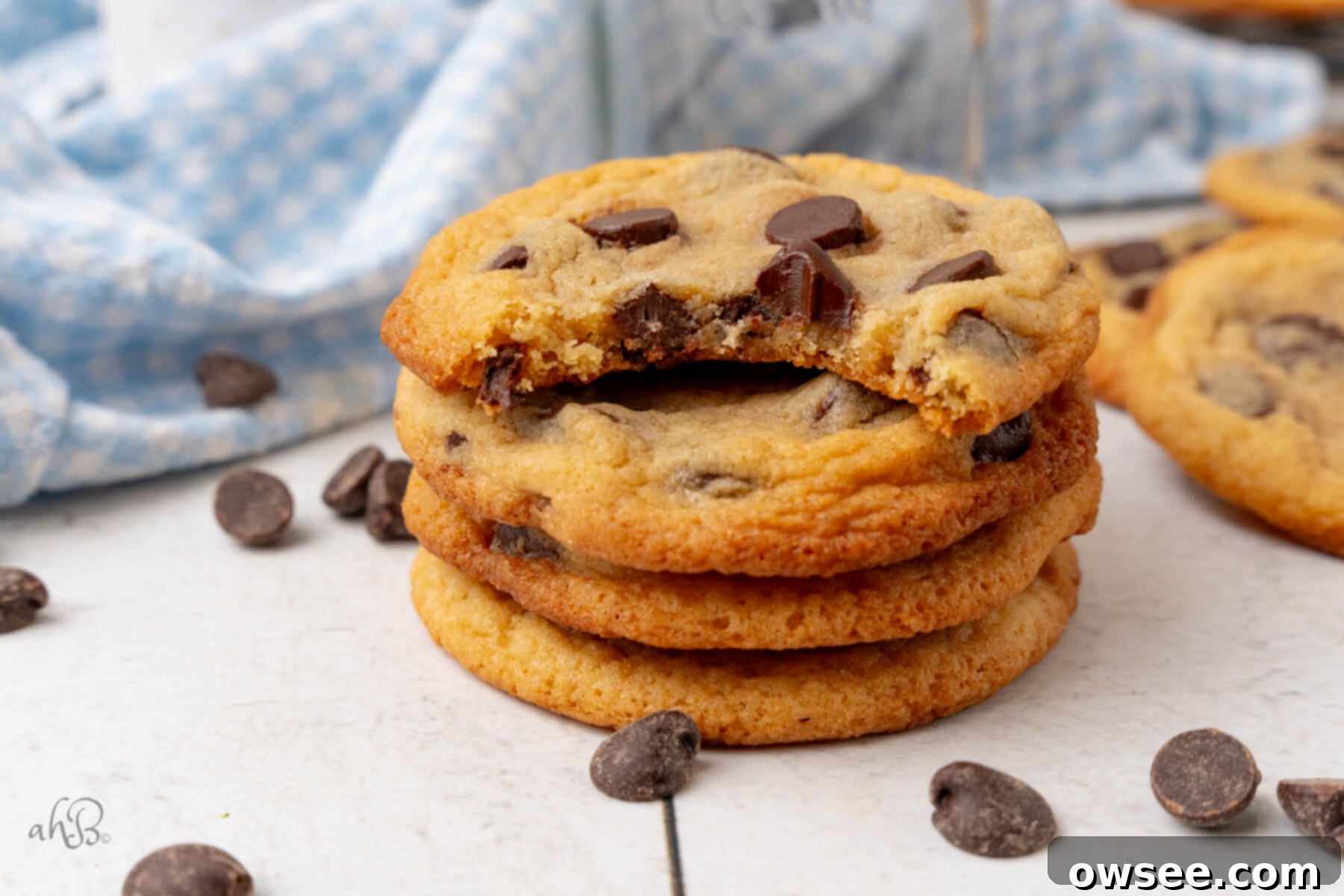 Amid a scattering of chocolate chips, a stack of four Malted Milk Cookies stands tall, with the top cookie having a perfect bite taken out, showcasing its moist, chewy interior.