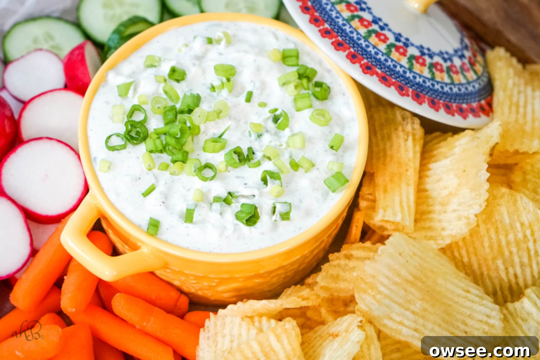 Green Onion Dip-7 A yellow dish of green onion dip is in the center of potato chips, and veggies with a lid off to the side.