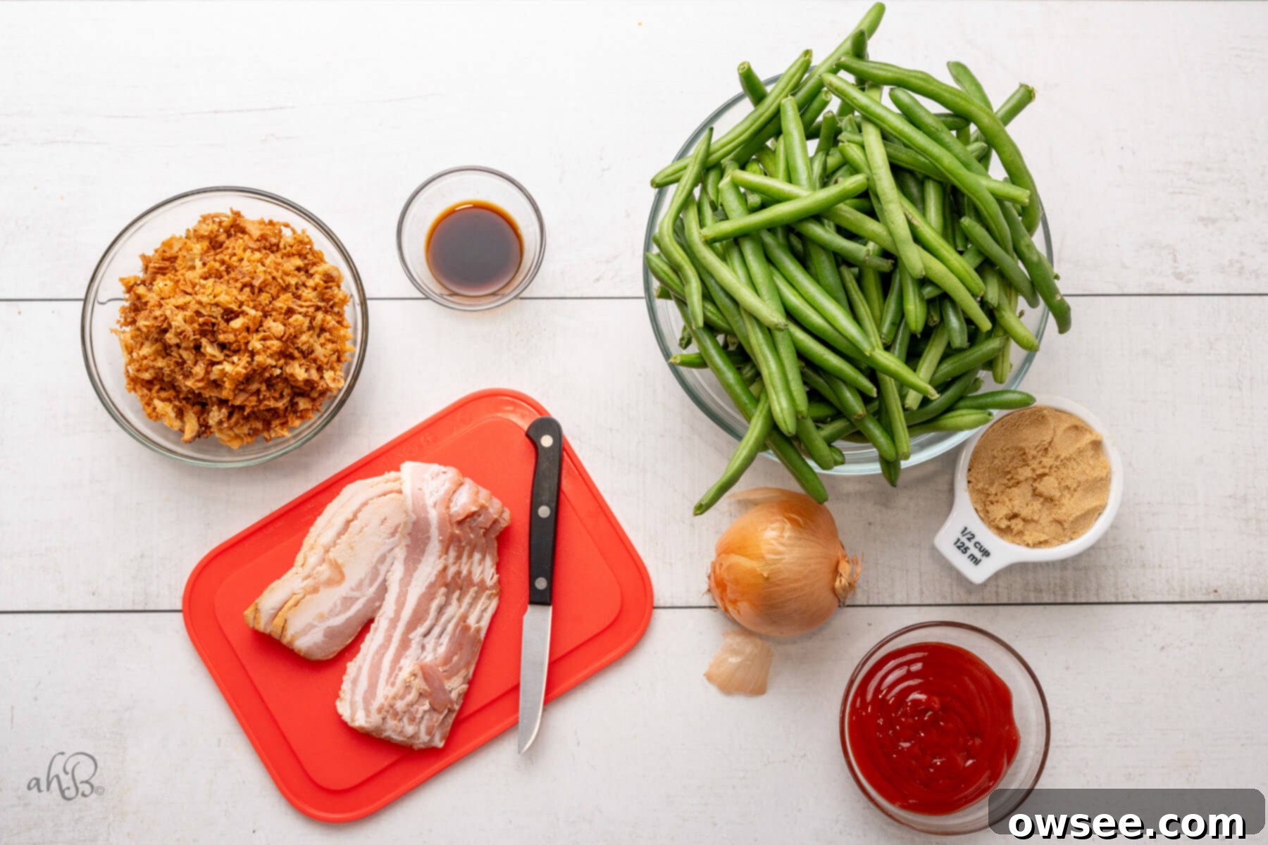 Smoky BBQ Green Bean Casserole 5 Slices of uncooked bacon, ketchup, a chopped onion, brown sugar, green beans, Worcestershire sauce, and French fried onions laid out on a wooden surface, ready for cooking.