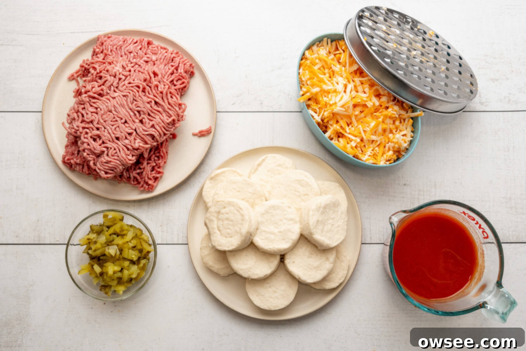 A flat lay showing all the simple ingredients for Sloppy Joe Biscuit Casserole: ground beef, canned biscuits, chopped dill pickles, shredded cheese, and a can of sloppy joe sauce.