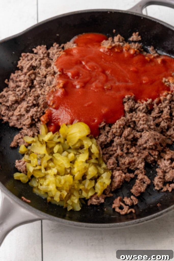 Sloppy joe sauce and chopped pickles being added to the browned ground beef in a skillet.