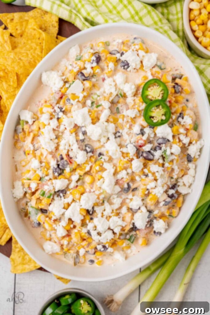 A white serving bowl full of fiesta corn dip topped with queso fresca cheese and fresh jalapeno pepper slices. The dip is creamy, colorful, and garnished for serving.