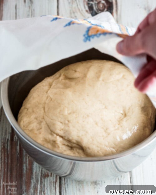 Homemade oatmeal dinner roll dough is resting in a large oiled bowl, covered with plastic wrap for its first rise.