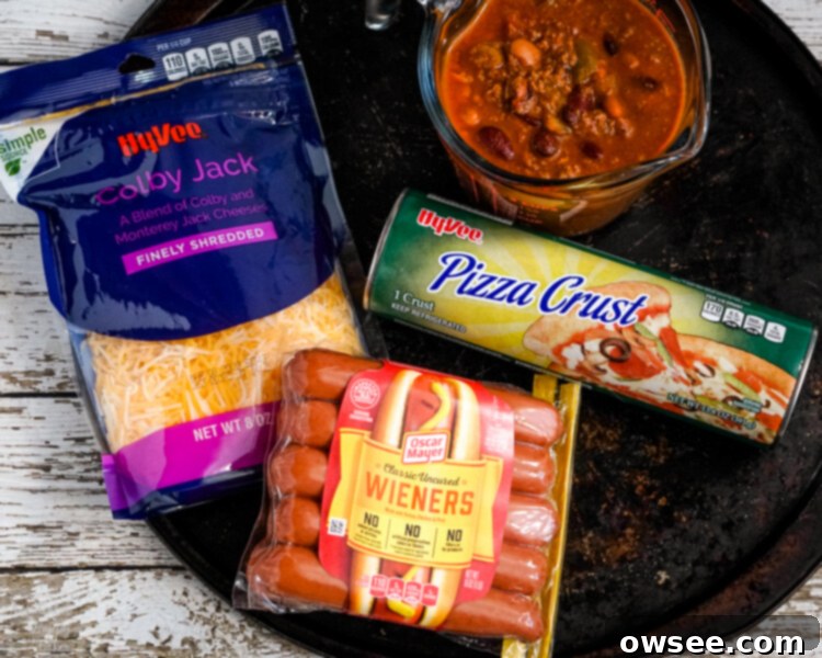 Chili Dog Pizza Fusion 3 Ingredients for Chili Dog Pizza laid out: shredded cheese, sliced hot dogs, a bowl of chili, and a pizza crust on a pizza pan.