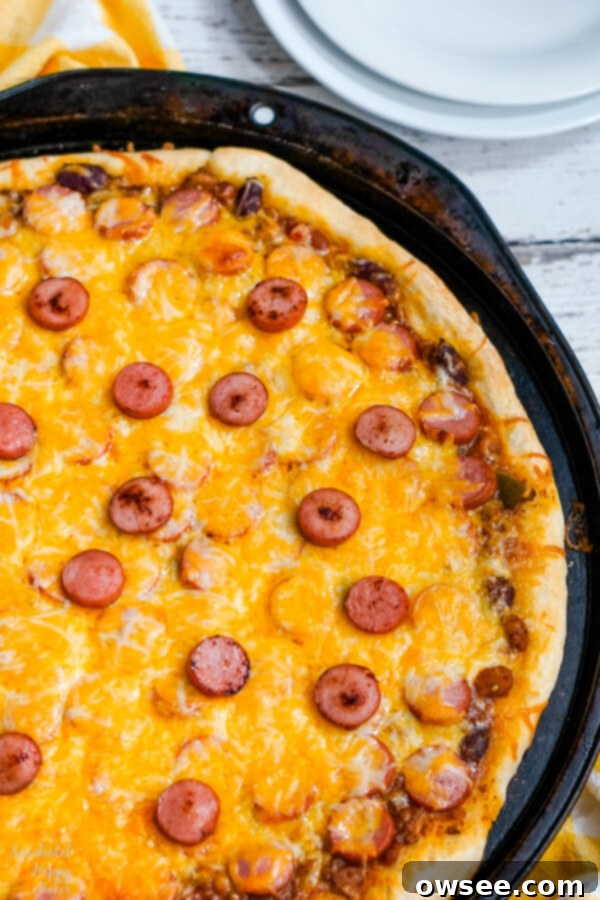 The Chili Dog Pizza is generously topped with shredded cheddar cheese, covering the hot dogs and chili, ready for baking.