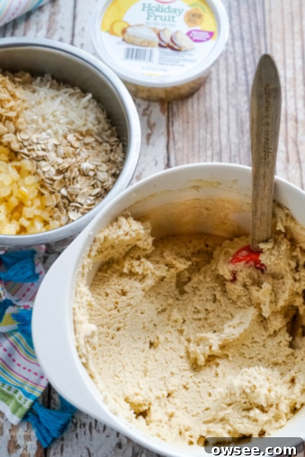 A pristine white bowl of creamy cookie dough awaits next to another bowl brimming with a colorful assortment of candied lemon peel, rolled oats, shredded coconut, and crispy rice cereal, ready for the next step in preparing Lemon Cowboy Cookies.