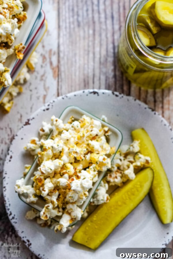 A bowl of freshly popped popcorn, generously seasoned with dill and spices, sits invitingly next to an opened jar of vibrant green dill pickles.