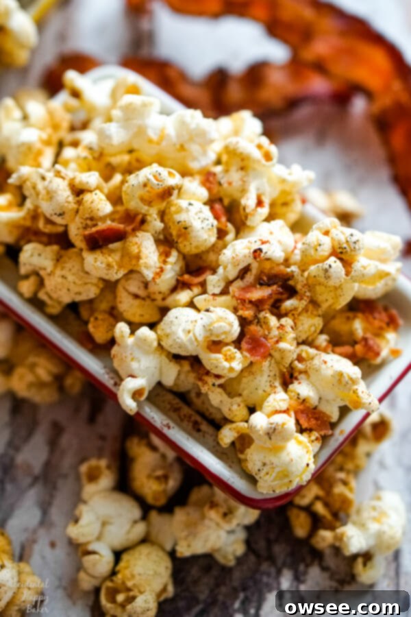 A close-up overhead view of a bowl of fluffy popcorn, generously topped with crispy bacon pieces and a sprinkle of visible seasonings like red paprika and black pepper.