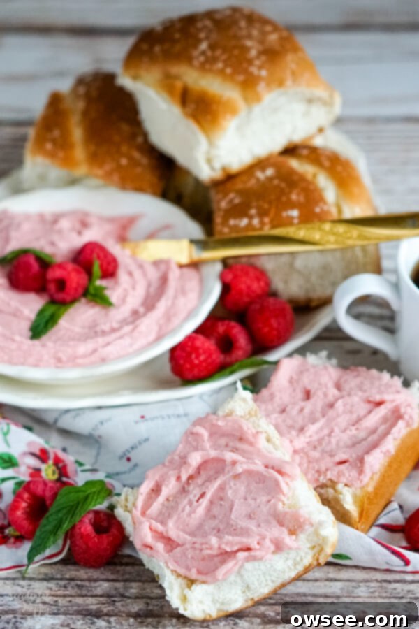 A split dinner roll generously slathered with creamy raspberry honey butter, with a serving platter of warm rolls in the background.