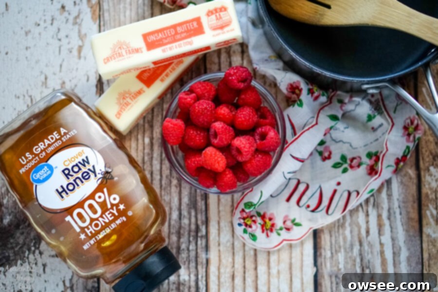 Three core ingredients for raspberry honey butter: fresh red raspberries, a jar of golden honey, and a stick of unsalted butter, arranged next to a small saucepan.