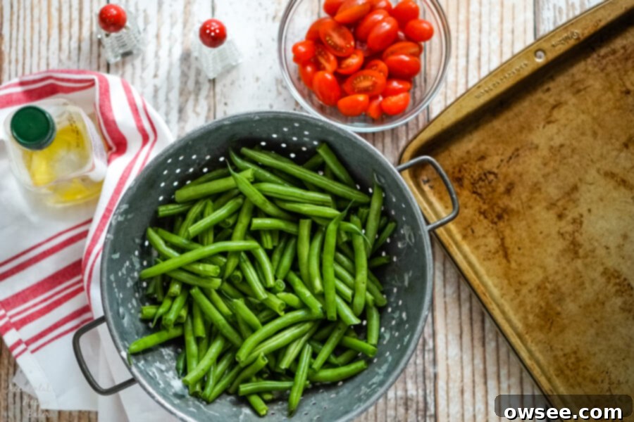 Green beans in a metal colander, next to a bowl of tomatoes, olive oil, salt and pepper.