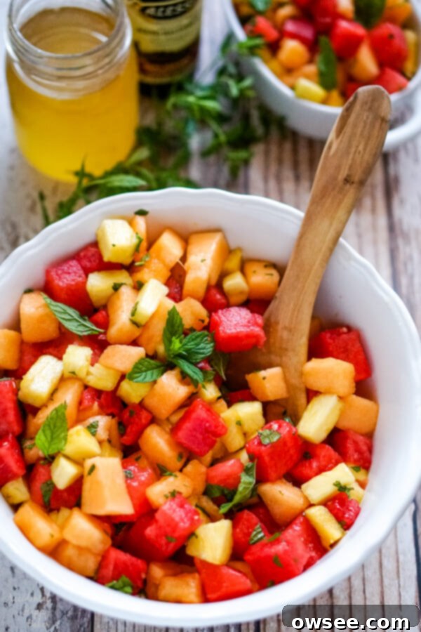 A white bowl of the freshly prepared watermelon fruit salad, garnished with mint, sitting next to a small jar of honey and a bottle of balsamic vinegar, highlighting the dressing ingredients.