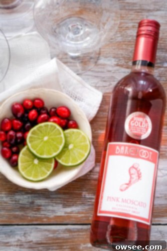Sparkling Cranberry Moscato Punch 3 A bottle of sparkling pink moscato wine stands beside a bowl overflowing with fresh cranberries and a plate of bright green sliced limes, highlighting the simple ingredients for the punch.