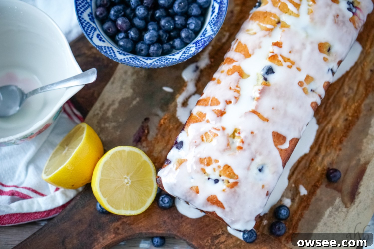 Zesty Lemon Blueberry Bread Drizzled with Buttermilk Glaze 7 A beautifully glazed lemon blueberry buttermilk bread on a wooden board, flanked by fresh lemons and blueberries, ready to be sliced and served.