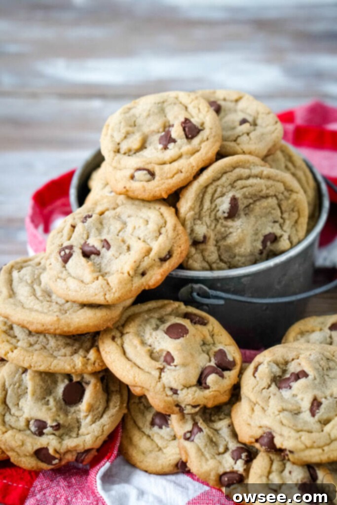 A large stack of golden brown, soft chocolate chip cookies next to a metal bucket brimming with more cookies, highlighting the generous yield of this big batch recipe.