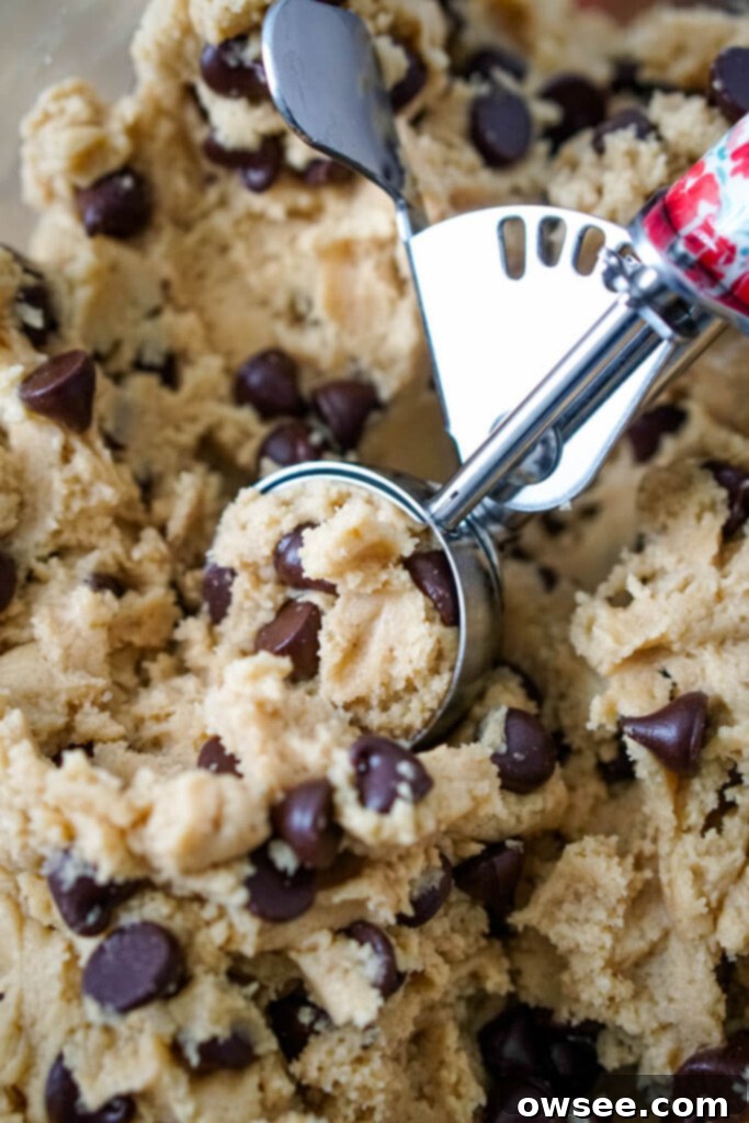 A large stainless steel mixing bowl filled with fluffy, golden chocolate chip cookie dough, with a metal cookie scoop resting on top, ready for action.