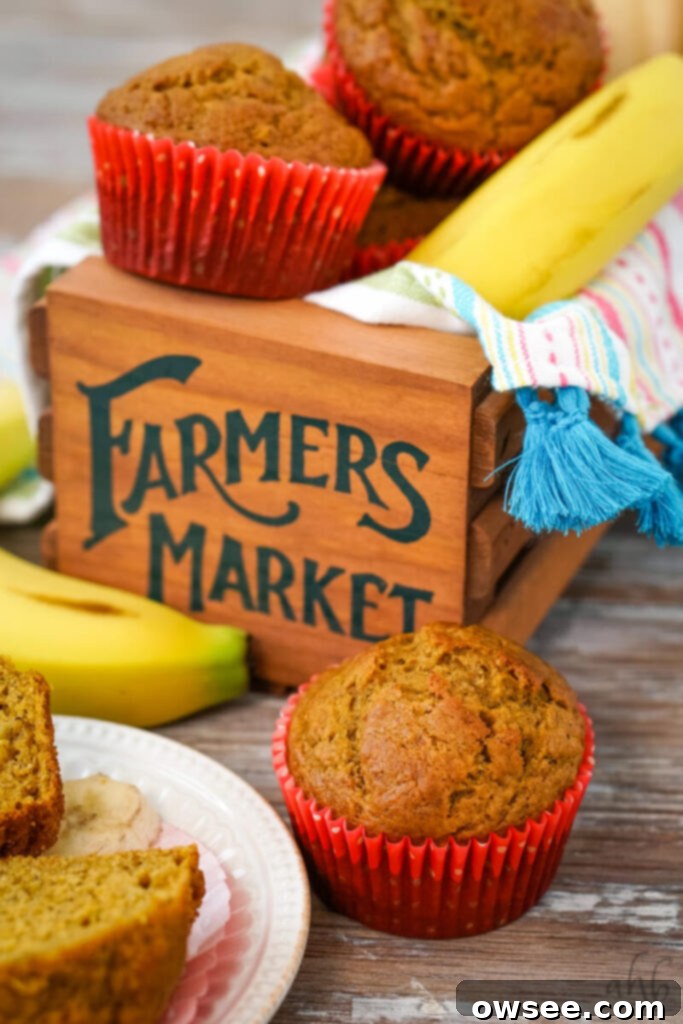 Golden Spiced Banana Pumpkin Muffins 3 A close-up shot of a pumpkin banana muffin in a red paper liner next to a sliced-open muffin revealing its moist interior, with a ripe banana in the background. Two more muffins and a banana are visible on a wooden 'Farmers Market' box.