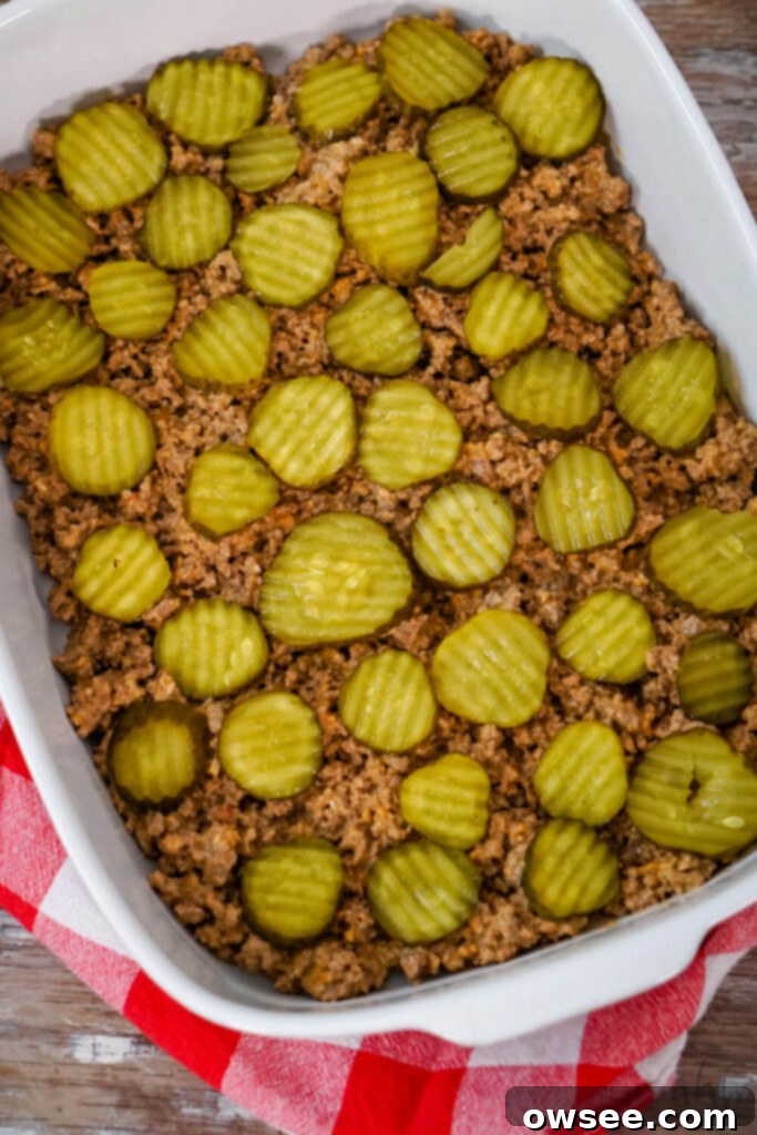 Big Mac Tater Tot Bake 8 Browned ground beef is evenly spread in the bottom of a white baking dish. Pickle slices are evenly placed on top.