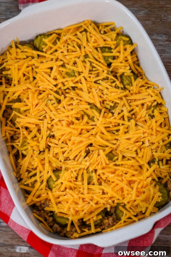 Big Mac Tater Tot Bake 9 Ground beef and pickles are topped with shredded cheddar cheese in a white baking dish.